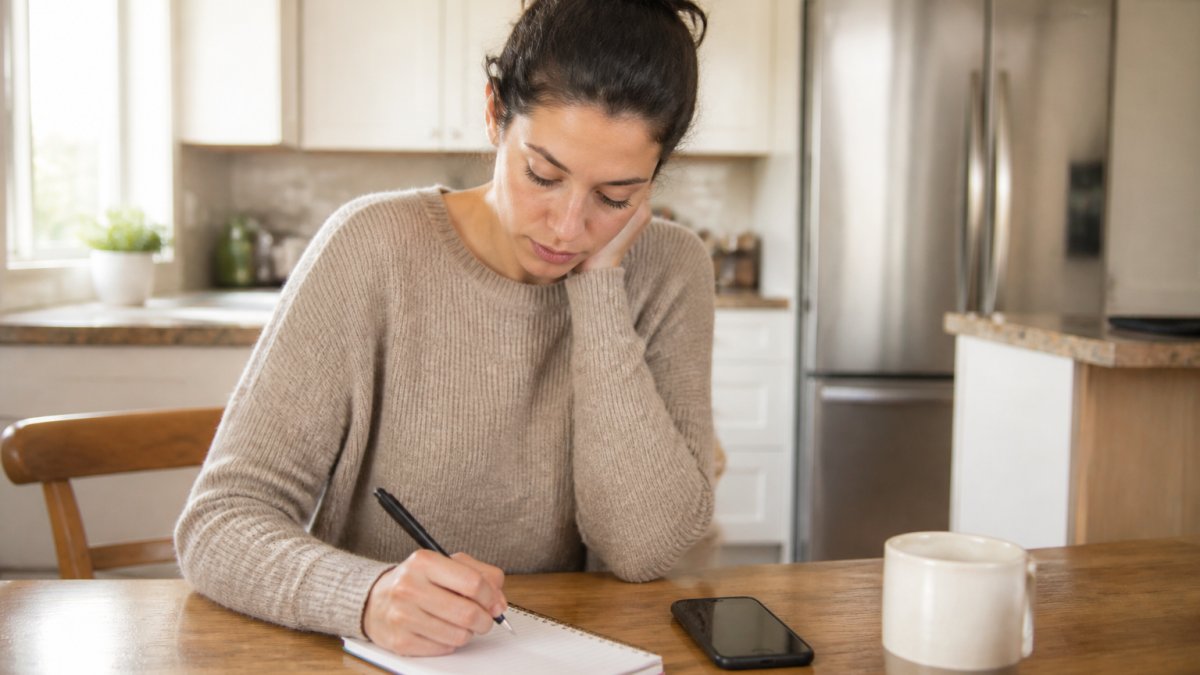 A person sitting at a kitchen table with a notepad and phone, jotting down questions before calling a dental clinic, warm natural light